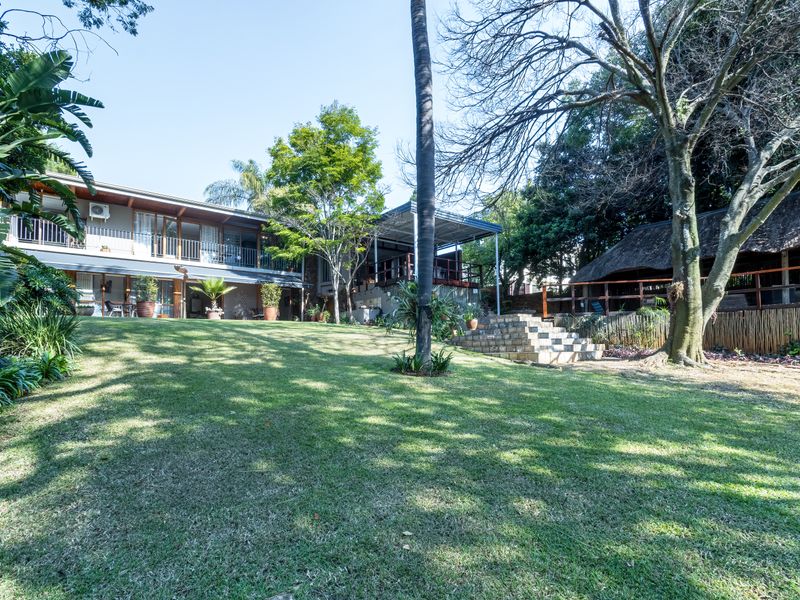 Spacious rear garden featuring a large lawn, mature trees, stone steps, and a multi-level dwelling with a balcony and separate thatched outdoor entertainment area.