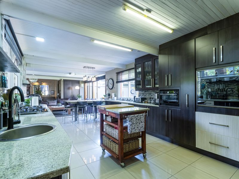 Spacious open-plan kitchen and living area featuring modern dark cabinetry, granite countertops, built-in appliances, and tiled flooring under a timber panelled ceiling.