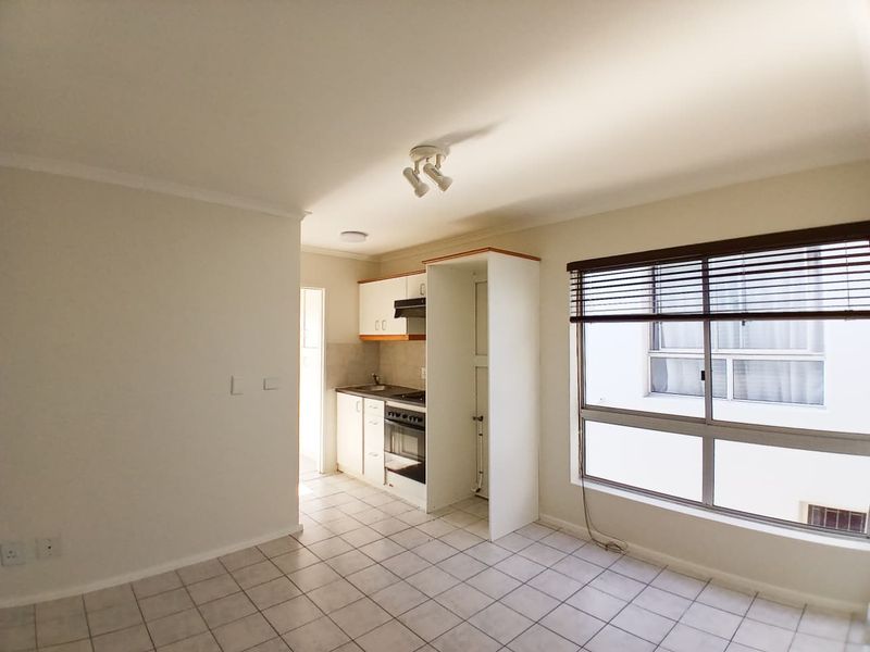 An open-plan kitchen area featuring tiled flooring, built-in cabinetry, an oven, and a hob, adjacent to a window with venetian blinds.