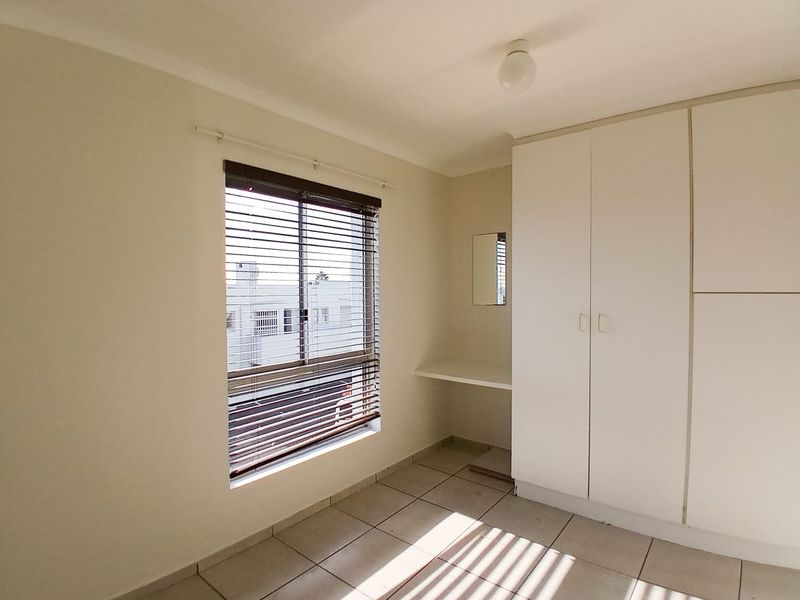 A bright, empty bedroom featuring tiled flooring, built-in cupboards, a small vanity area, and venetian blinds.