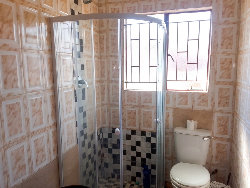 A compact bathroom featuring a corner shower enclosure, standard toilet, and dated wall tiling with security bars on the window.