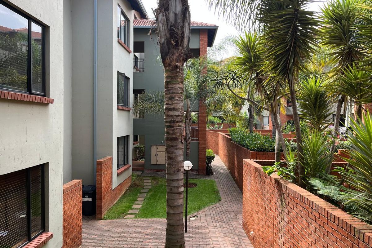 A well-maintained apartment complex exterior featuring brick boundary walls, paved walkways, and established garden landscaping between building blocks.