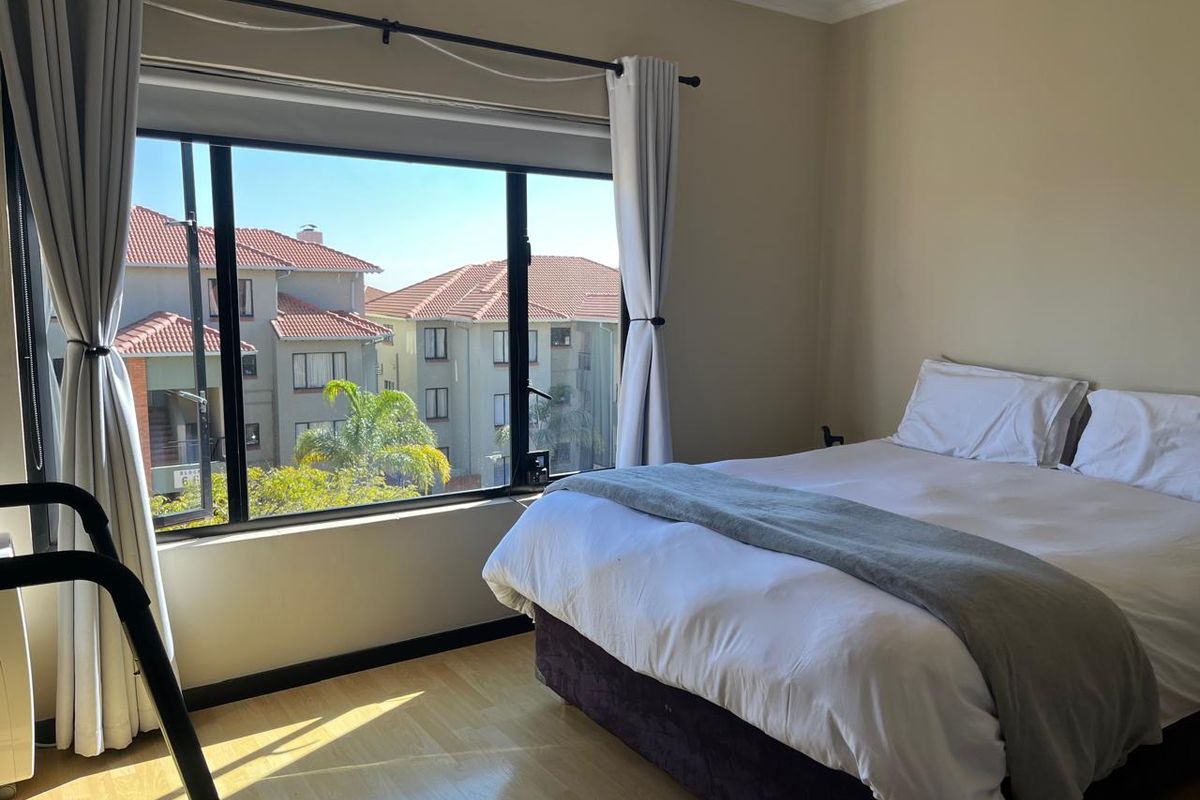 A bright bedroom featuring laminate flooring and a large window with views of the surrounding apartment complex.