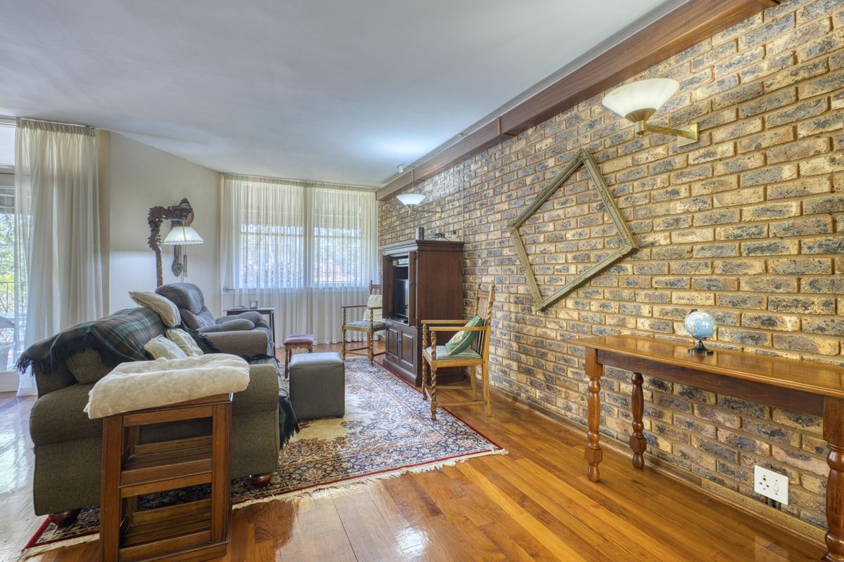 A cozy living room featuring a prominent exposed brick wall, polished hardwood flooring, and natural light streaming through large windows with sheer curtains.