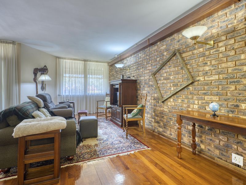A cozy living room featuring a prominent exposed brick wall, polished hardwood flooring, and natural light streaming through large windows with sheer curtains.
