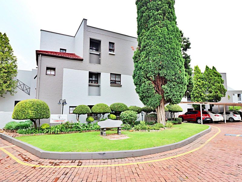 Front exterior view of the townhouse complex featuring a paved driveway, landscaped garden area, and covered parking bays.