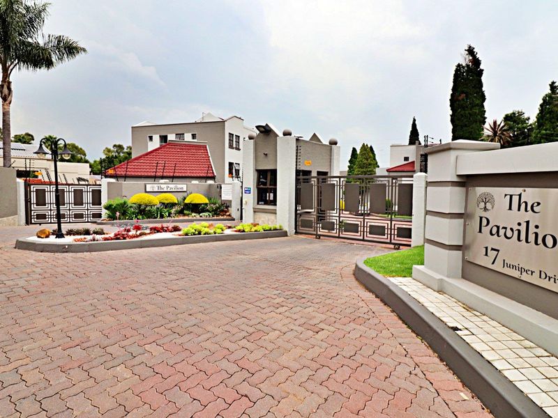 The secure entrance to The Pavilion complex at 17 Juniper Drive, featuring a paved driveway, guard house, and well-maintained landscaped garden beds.