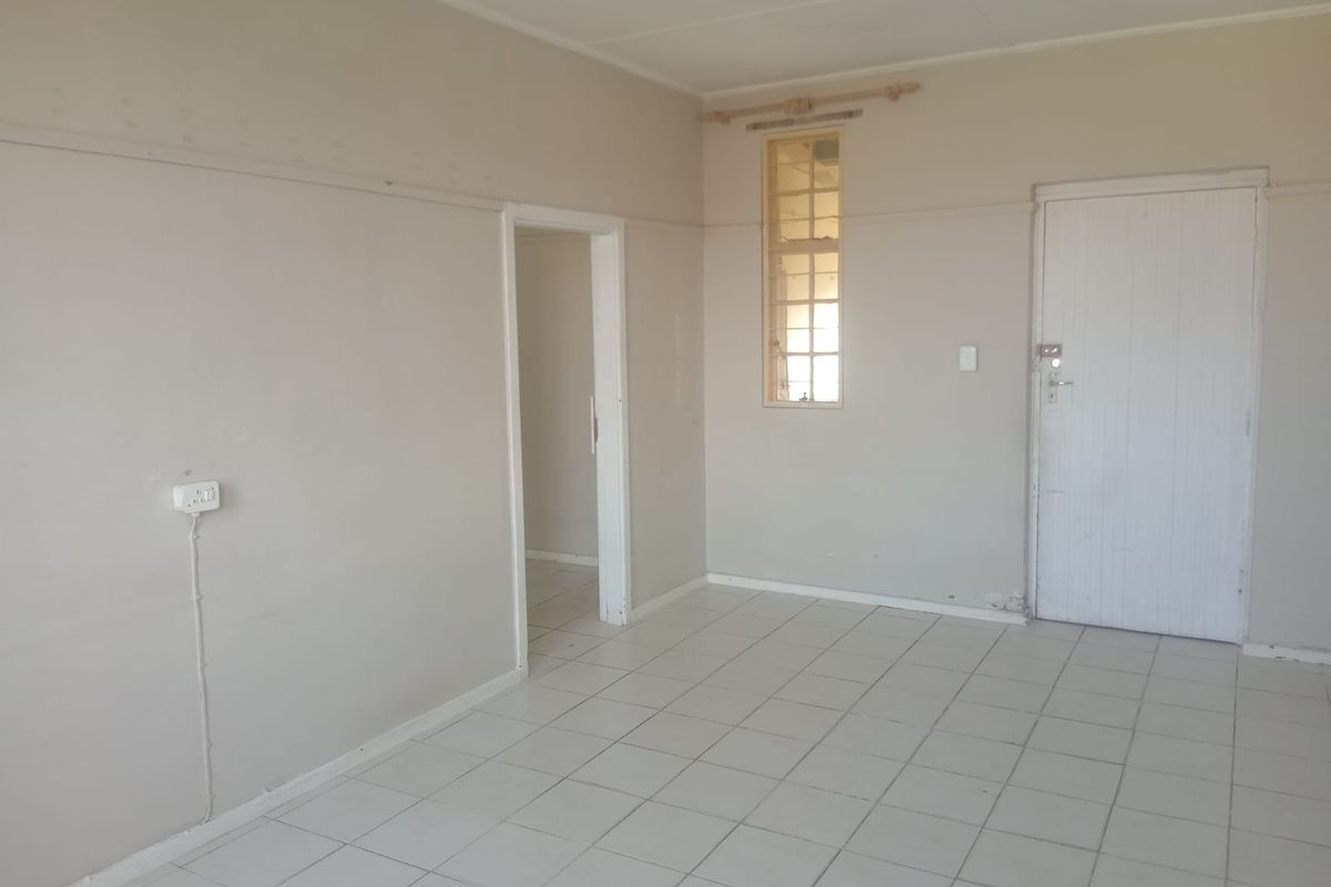 An empty, neutrally painted living area featuring ceramic floor tiles, a single steel-framed window, and a standard interior door.