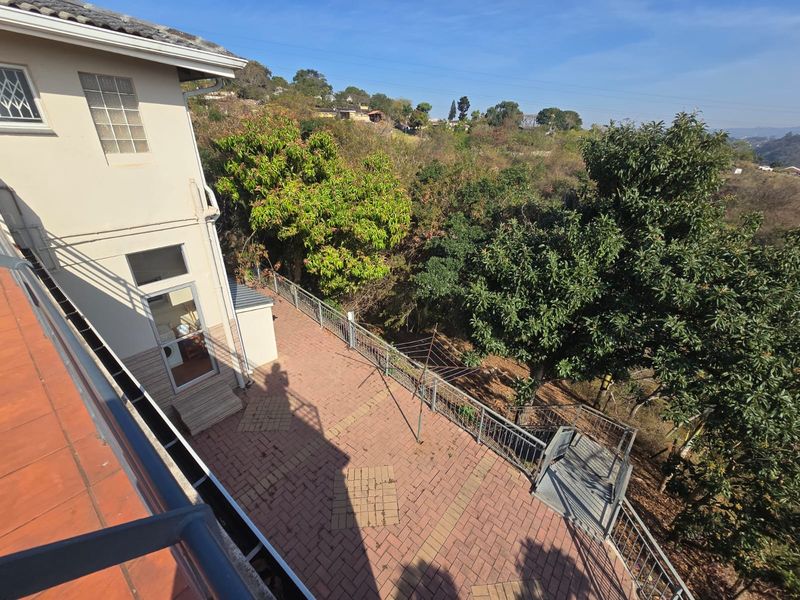 An elevated view of a paved rear patio area featuring metal railings, an external staircase, and access to the garden with mature trees.