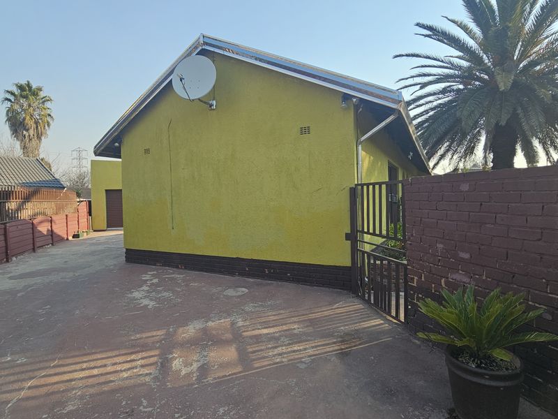 Rear exterior view of the property featuring a yellow facade, concrete driveway, and brick boundary wall with a metal security gate.