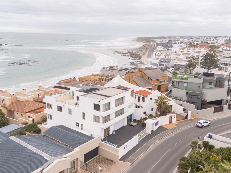 Aerial view of a multi-story coastal property featuring flat roofs, balconies, and a paved driveway, situated in a prime seaside residential area.