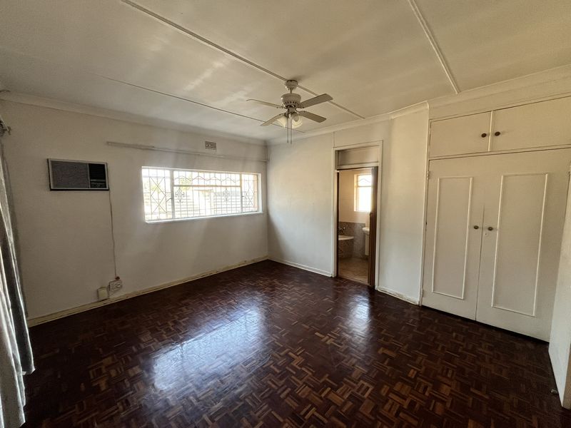 A spacious bedroom featuring classic parquet flooring, built-in cupboards, a ceiling fan, and an integrated air conditioning unit, with an adjoining bathroom.