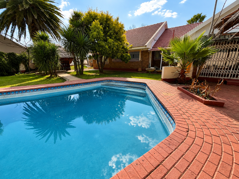 A backyard pool area featuring a swimming pool with brick paving, surrounded by established garden landscaping and a view of the house exterior.