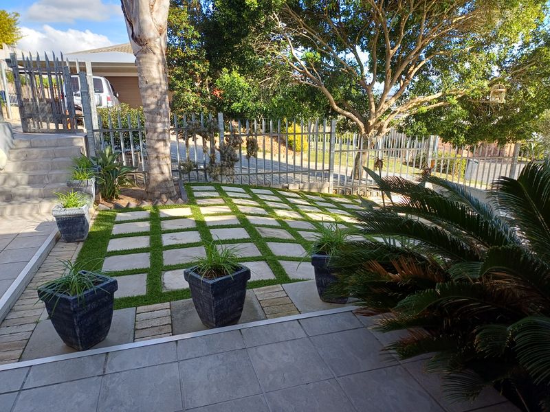 A well-maintained garden area featuring a paved grid walkway with artificial grass, metal security fencing, and mature trees.