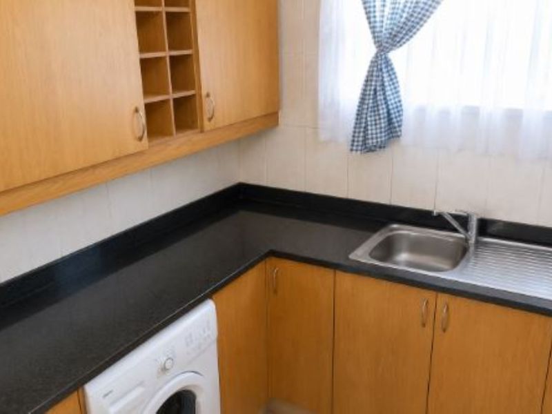A functional kitchen area featuring light wood-tone cabinetry, black countertops, a stainless steel sink, and a tiled splashback.