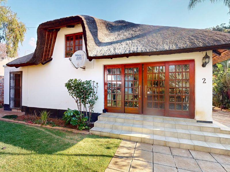 Front elevation of a cottage-style home featuring a traditional thatch roof, timber-framed glass doors, and tiled entrance steps set within a garden.