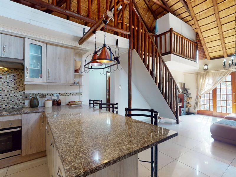 Open-plan kitchen and living area featuring granite countertops, a built-in oven, a unique thatched roof, and a wooden staircase leading to a mezzanine level.