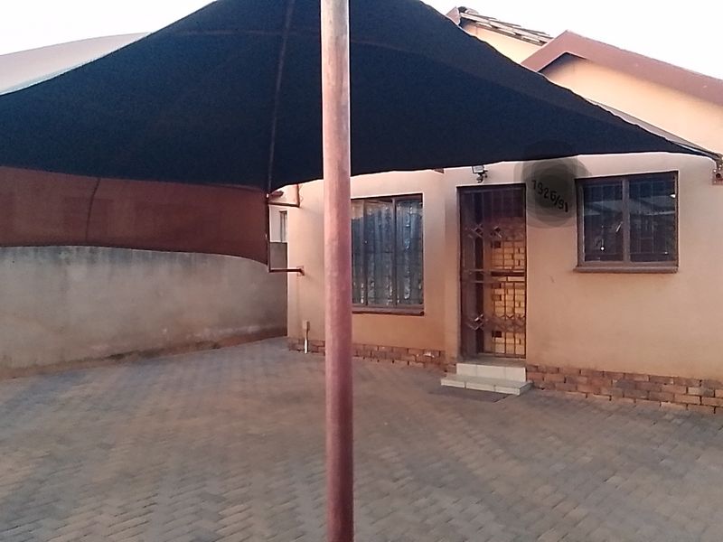 Front exterior view of the property featuring a paved driveway, a large shade sail, and secure entrance with burglar bars.