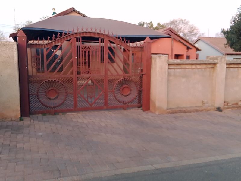 Front view of the property featuring a secure wrought iron gate, paved driveway, and perimeter walling with a carport structure.