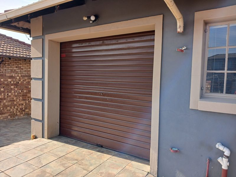 A view of a garage exterior featuring a brown roller shutter door, tiled driveway area, and adjacent window with security bars.