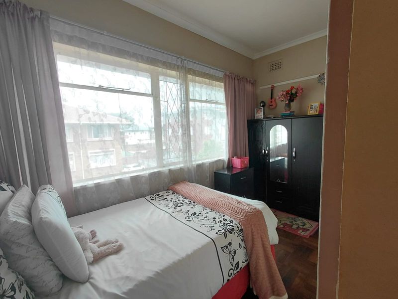 A bedroom featuring parquet flooring and a large window with security bars, providing natural light and a view of the surrounding apartment complex.