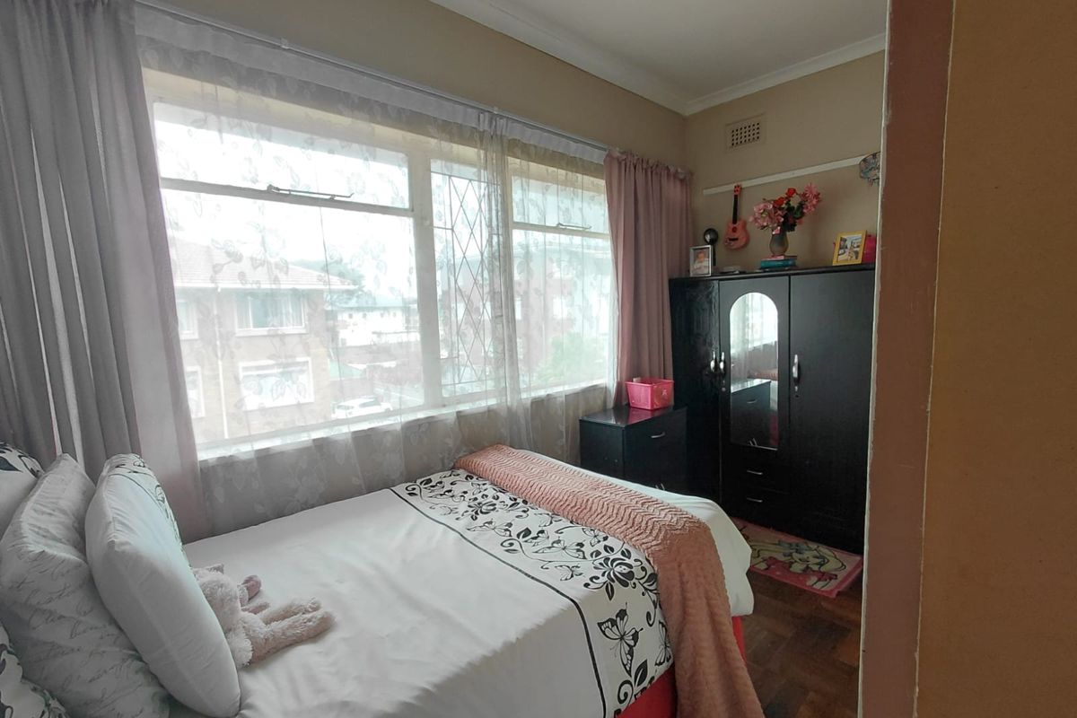 A bedroom featuring parquet flooring and a large window with security bars, providing natural light and a view of the surrounding apartment complex.