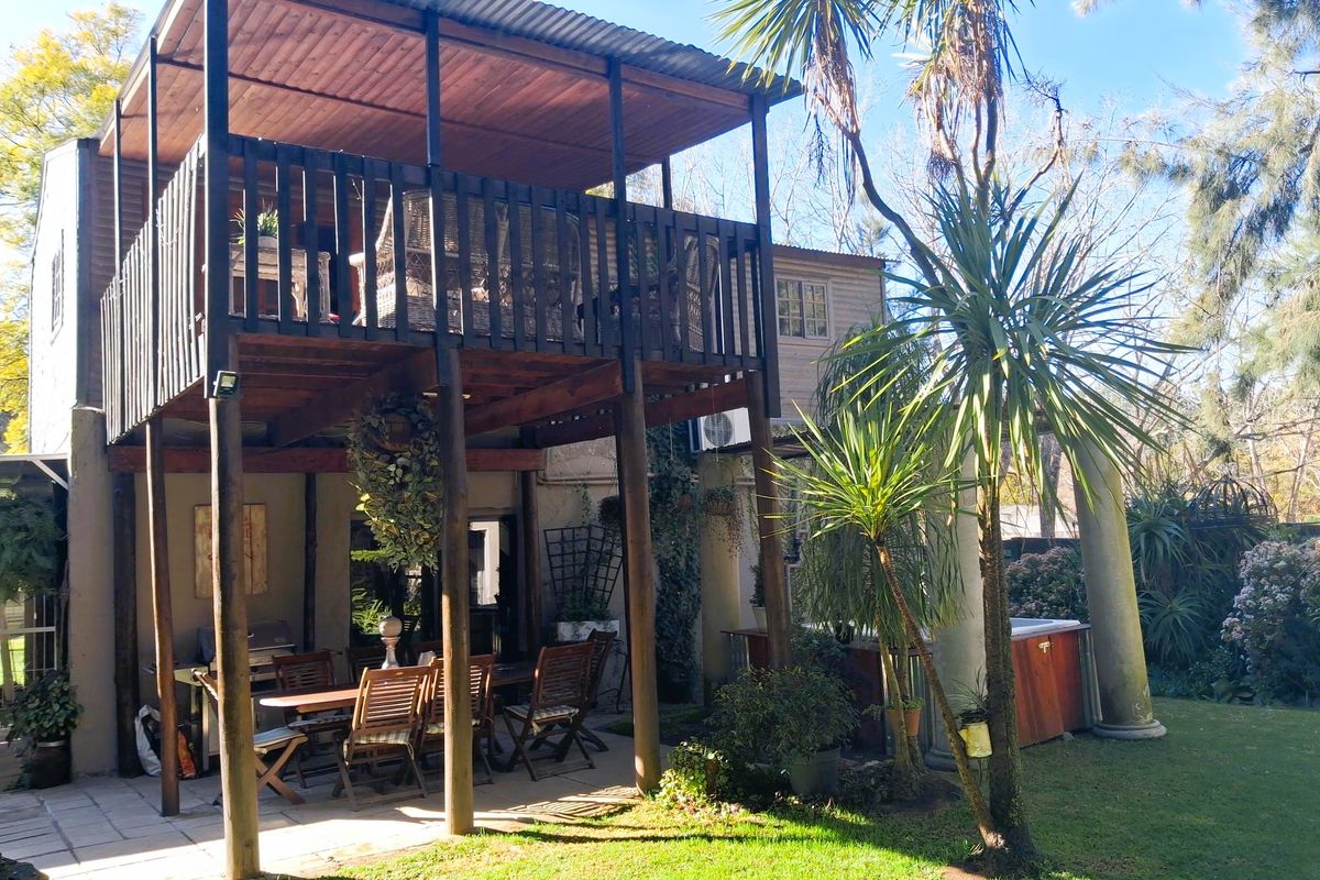 Rear exterior view featuring a two-level timber deck, covered ground-floor patio, paved entertainment area, and a built-in hot tub set in a garden.