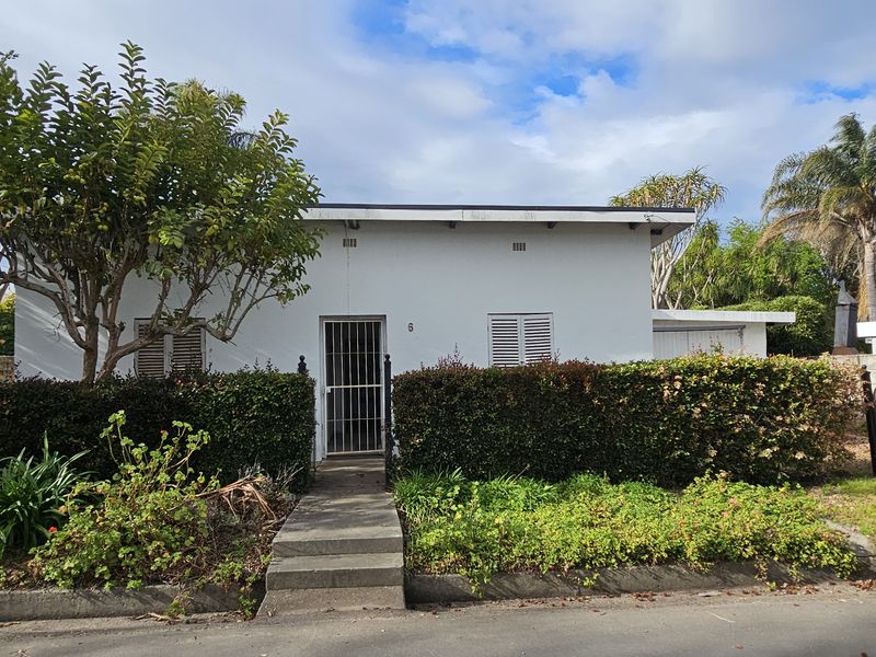 Front elevation of a single-story house featuring a security gate, concrete entrance steps, and a surrounding boundary wall with established garden greenery.