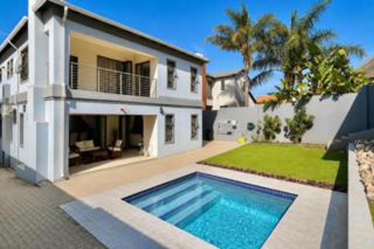 Rear view of a modern two-story house featuring a swimming pool, manicured lawn, paved patio area, and an upper-level balcony with stainless steel railings.