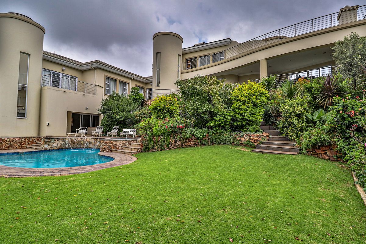 A spacious rear garden featuring a swimming pool with a water feature, stone retaining walls, landscaped greenery, and an elevated balcony with metal railings.