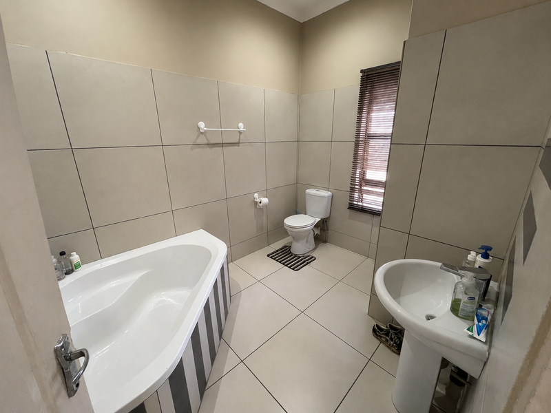 A functional bathroom featuring a corner bathtub, pedestal basin, toilet, and neutral floor-to-ceiling wall tiling with a window blind.