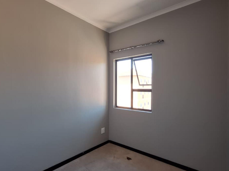 A clean, empty bedroom featuring neutral grey walls, a casement window with a curtain rod, and carpeted flooring.