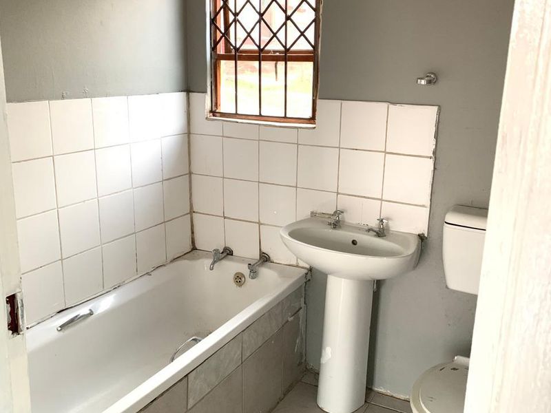 A dated bathroom featuring a standard bathtub, pedestal basin, and toilet, with tiled splashbacks and security bars on the window.