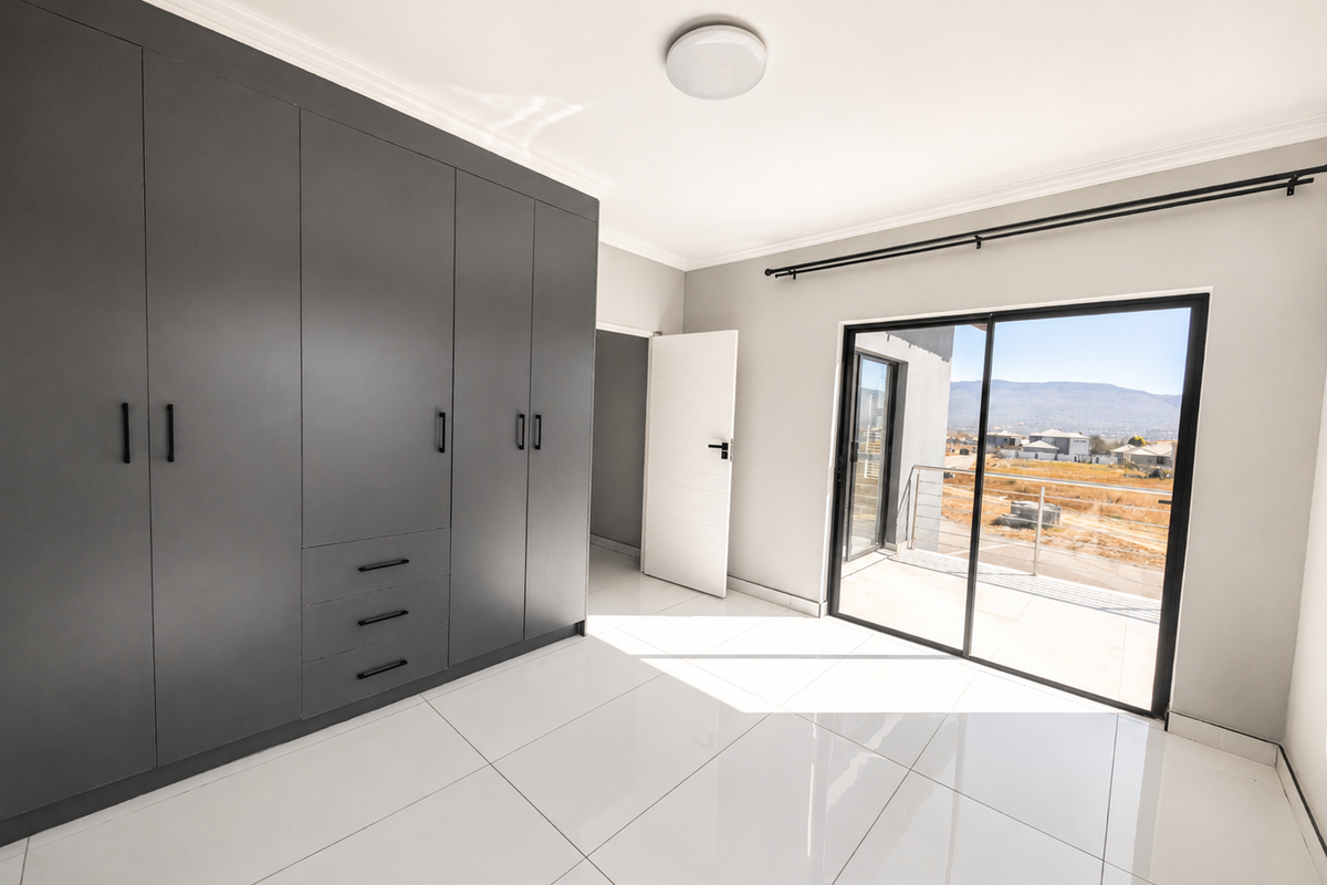 A bright, modern bedroom featuring sleek dark grey built-in cupboards, polished white floor tiles, and a sliding glass door leading to an outdoor balcony.