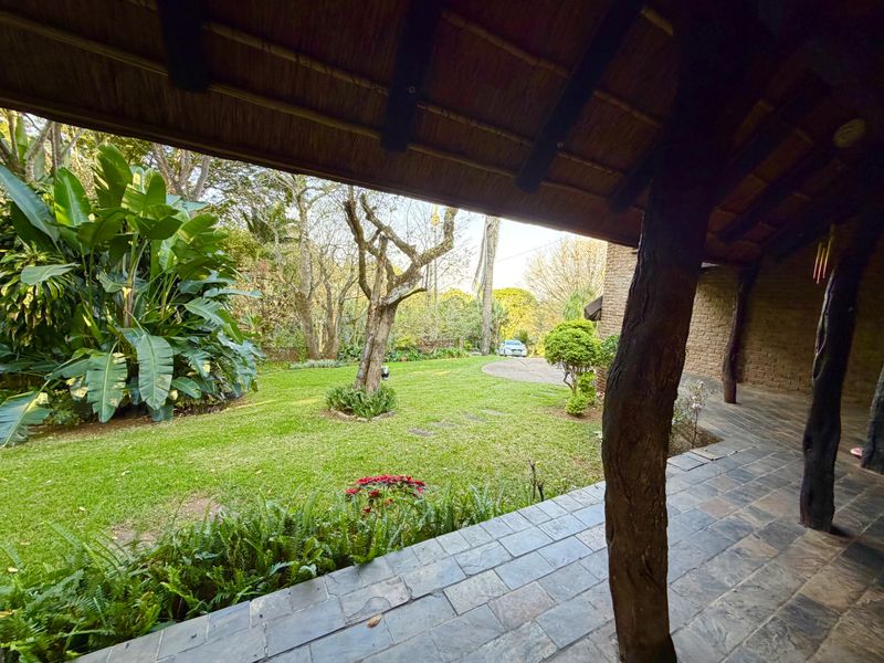 A covered patio featuring a rustic thatched roof, natural timber columns, and slate flooring, overlooking a lush, well-maintained garden.