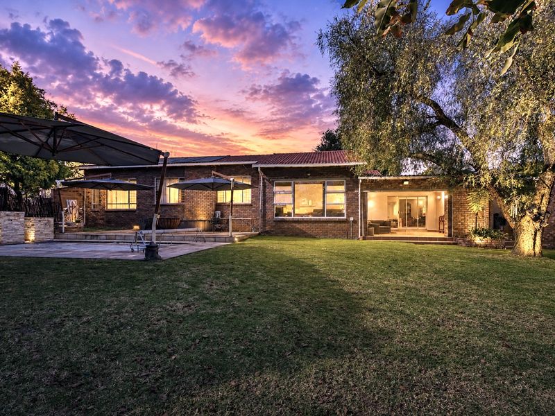 A beautifully lit rear exterior view of the house featuring a spacious paved patio, well-maintained garden, and warm evening lighting.