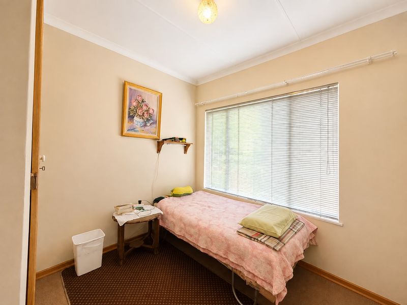 A compact bedroom featuring neutral wall tones, carpeted flooring, and large windows fitted with venetian blinds.