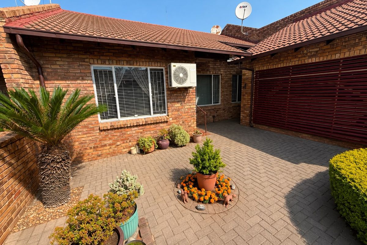 A well-maintained townhouse front entrance featuring a paved courtyard, face brick exterior, and integrated air conditioning unit.