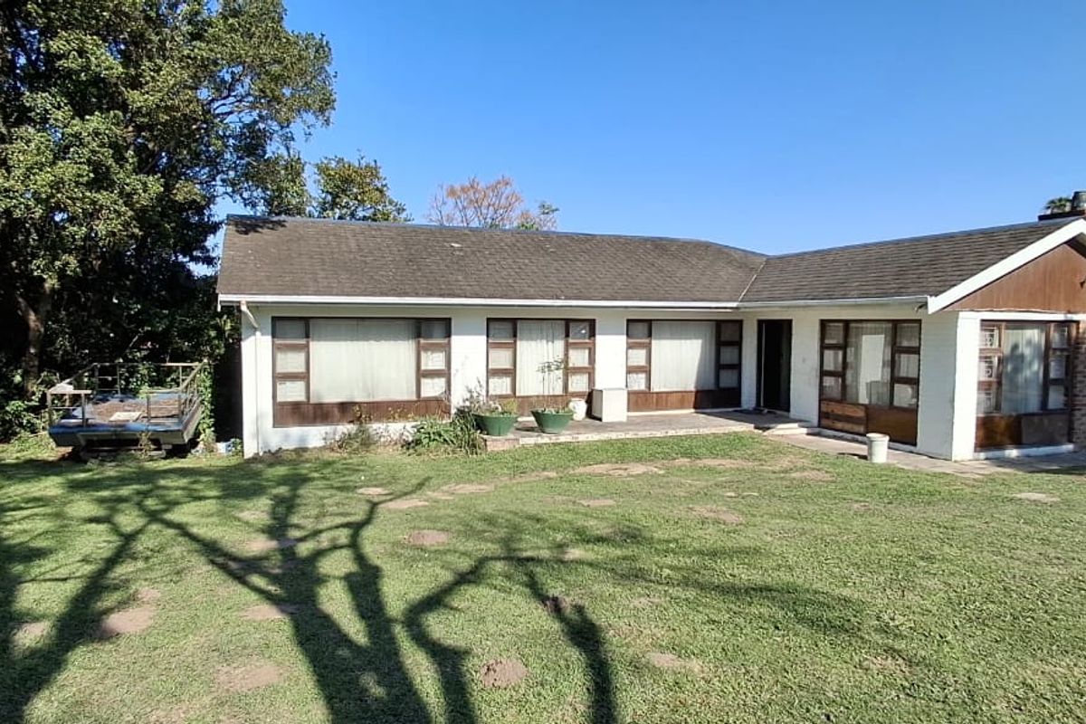 A rear view of a single-story residential house featuring a large lawn, paved patio area, and traditional brick and white-painted exterior finishes.