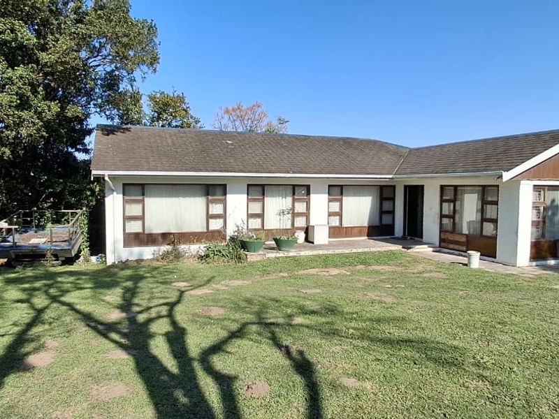 A rear view of a single-story residential house featuring a large lawn, paved patio area, and traditional brick and white-painted exterior finishes.