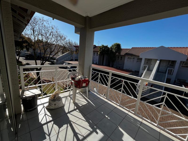 A spacious covered balcony featuring tiled flooring and a white metal balustrade, offering an open view of the apartment complex grounds.