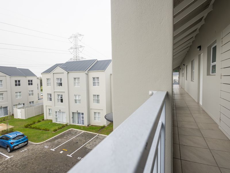 A covered exterior walkway or balcony featuring tiled flooring, a metal balustrade, and exposed rafters, overlooking the apartment complex grounds.