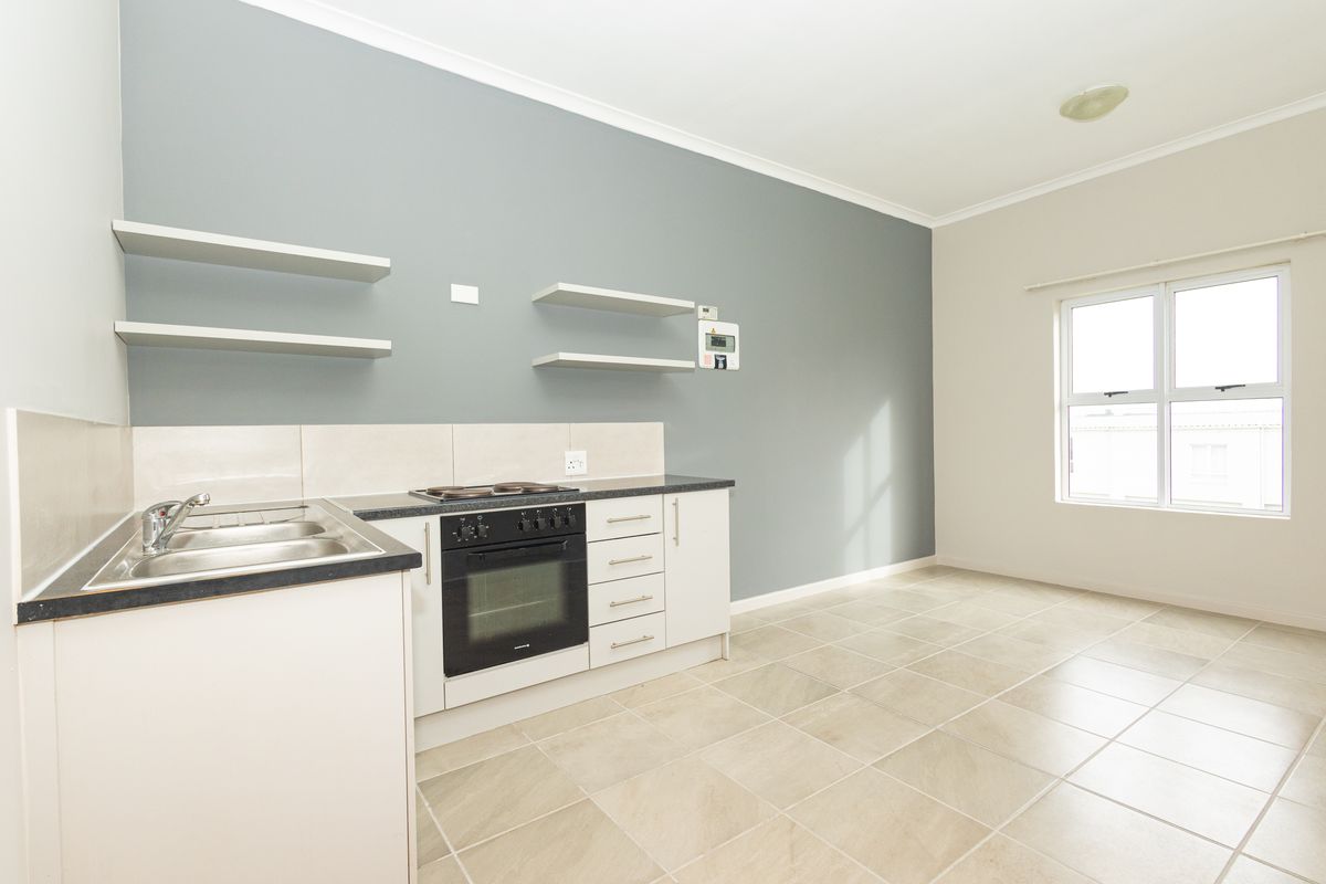 A clean, empty kitchen featuring built-in oven, electric hob, stainless steel sink, and floating shelves with neutral tiled flooring.