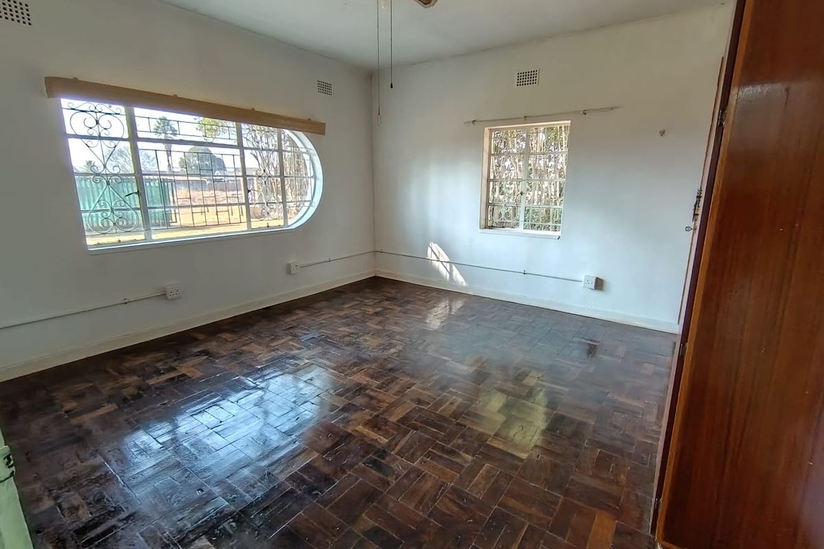 A spacious, empty bedroom featuring original parquet flooring, a ceiling fan, and large windows with security bars.
