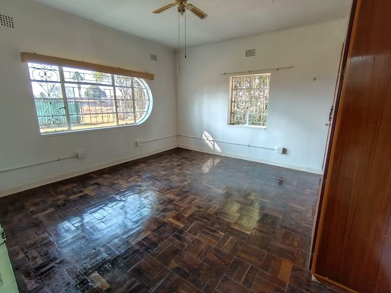 A spacious, empty bedroom featuring original parquet flooring, a ceiling fan, and large windows with security bars.