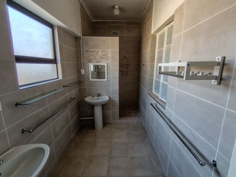 A functional bathroom featuring a pedestal washbasin, walk-in shower, and neutral ceramic tiling throughout with integrated towel rails and glass shelving.