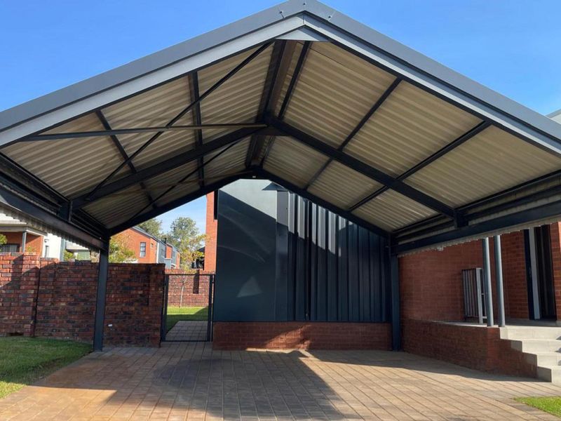 A modern, well-maintained steel carport with a corrugated roof, paved driveway, and brick boundary wall, providing covered parking space.