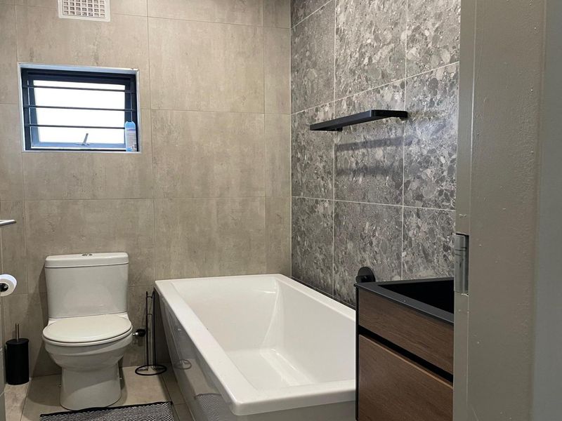 Modern bathroom featuring a built-in bathtub, vanity cabinet, and contemporary grey tiled walls with a small window.