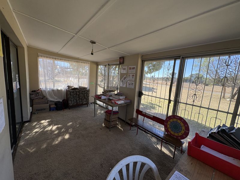 A sunlit living area featuring large sliding glass doors with security gates, carpeted flooring, and an exposed beam ceiling, offering views of the exterior.