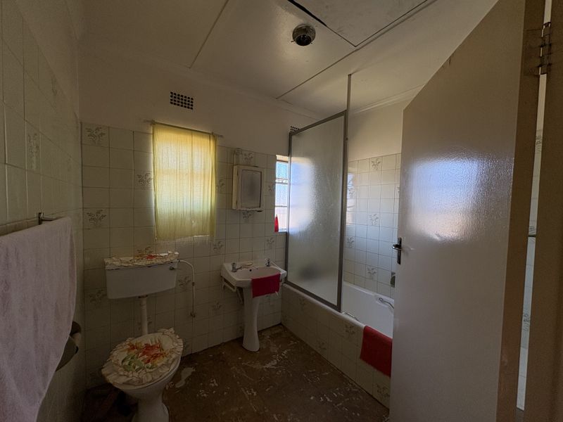 A dated bathroom featuring a pedestal basin, bathtub with shower screen, and toilet, requiring modernization of finishes and flooring.